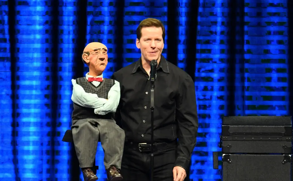 A ventriloquist and his puppet stand on stage with a blue backdrop.