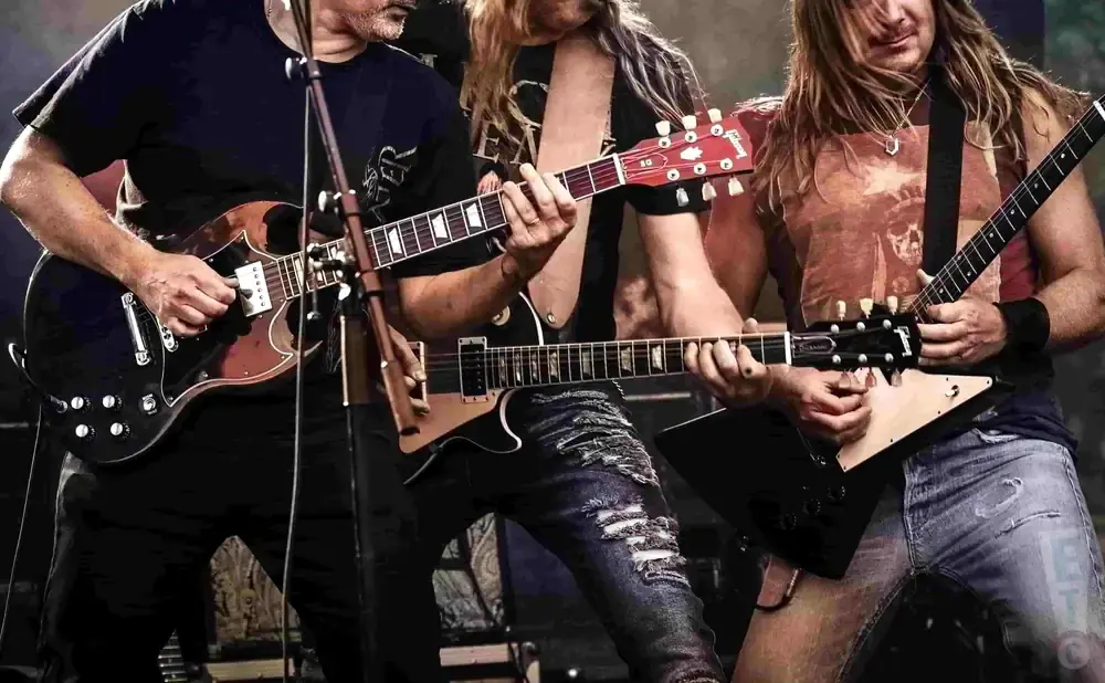 Three men with long hair playing electric guitars on stage.