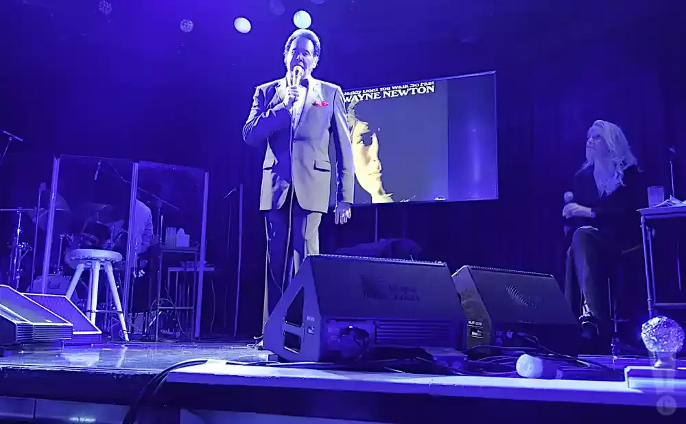 Wayne Newton on stage, holding a microphone, with a large screen displaying his album art behind him.