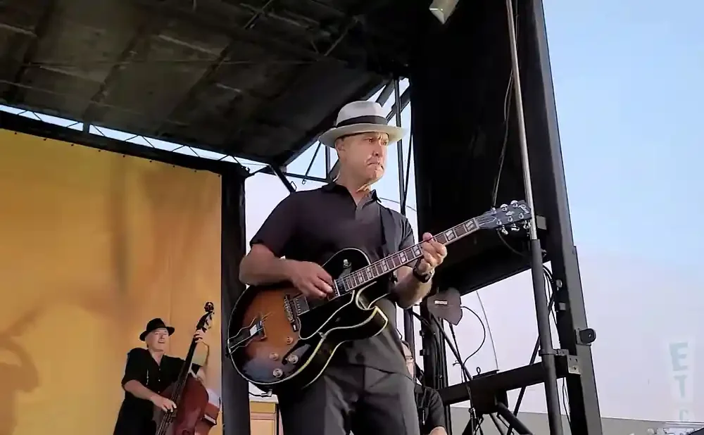 A man in a fedora plays a guitar on stage, with a bassist to his left.