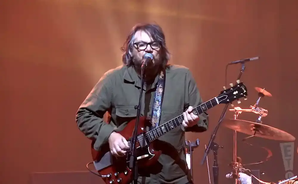 A guitarist in a green jacket plays a red electric guitar on stage with a microphone in front of him.