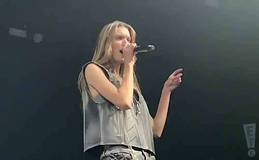 A woman sings into a microphone on stage, wearing a gray vest and patterned pants.
