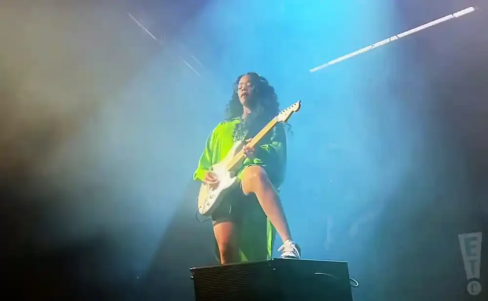 A woman in a bright green shirt plays a white electric guitar on stage under blue spotlights.