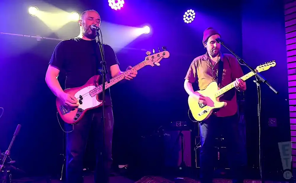 A band performs on a dimly lit stage with purple spotlights. Two guitarists are visible.
