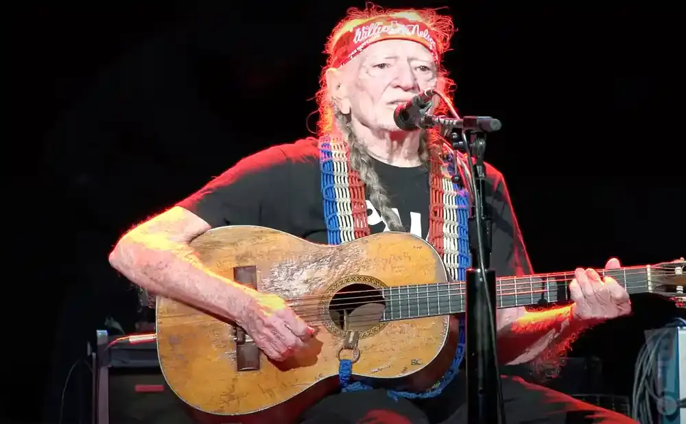 Willie Nelson plays his guitar, wearing a red bandana and a colorful beaded necklace, on stage.