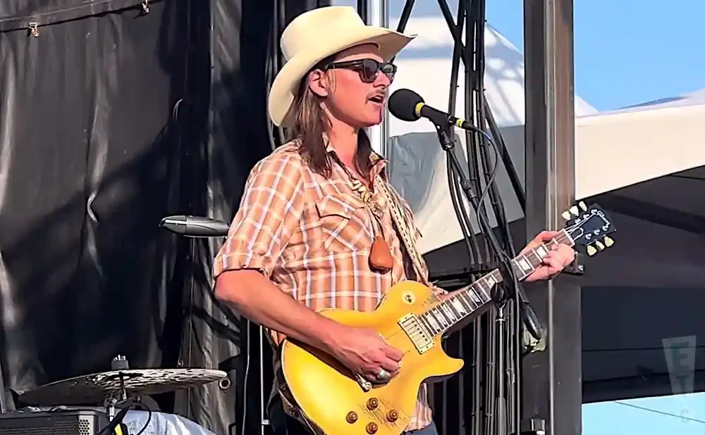 A man in a cowboy hat sings and plays a yellow electric guitar on stage.