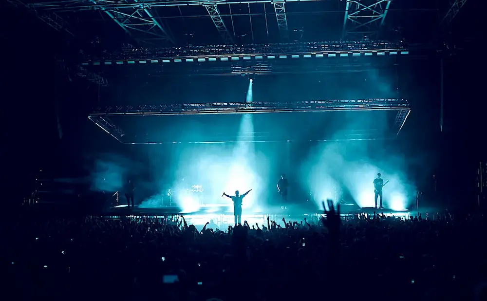 A band performs on a brightly lit stage for a cheering crowd, silhouetted against blue stage lights and smoke.