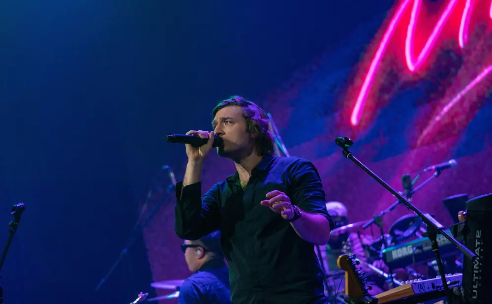 A male singer in a black shirt holds a microphone on stage, with pink and blue lights in the background.
