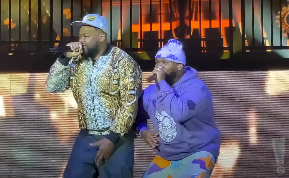 Two men performing on stage, one in a patterned jacket and light blue cap, the other in a purple hoodie and tie-dye beanie.
