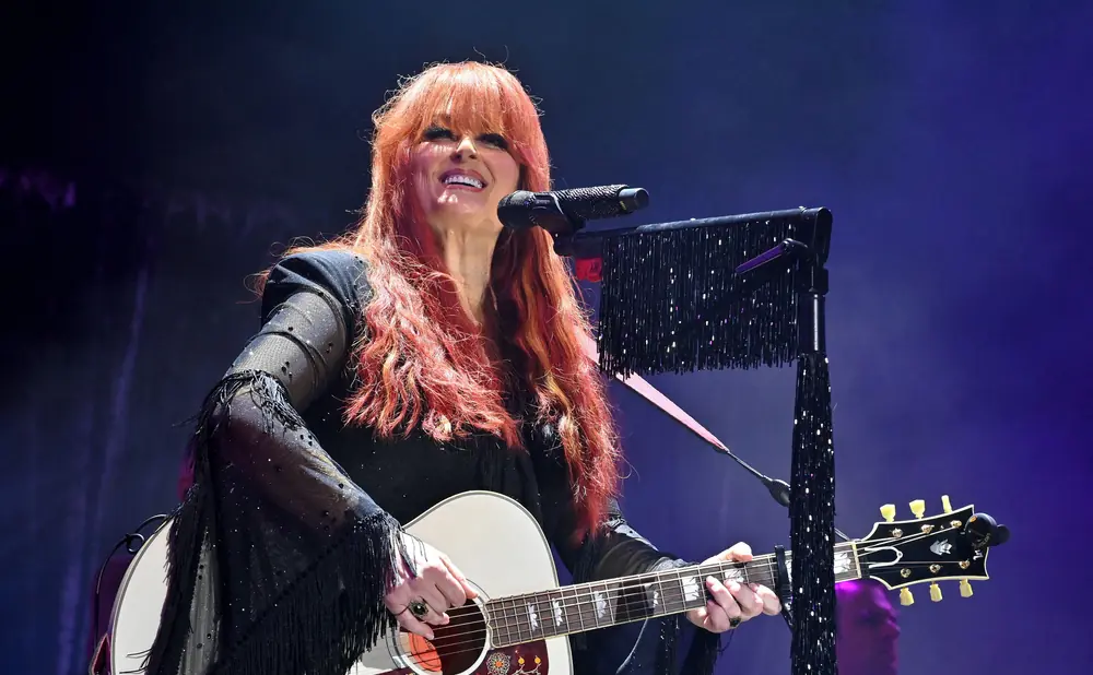 Singer Wynonna Judd on stage with a white acoustic guitar and red hair, wearing a black fringed dress.