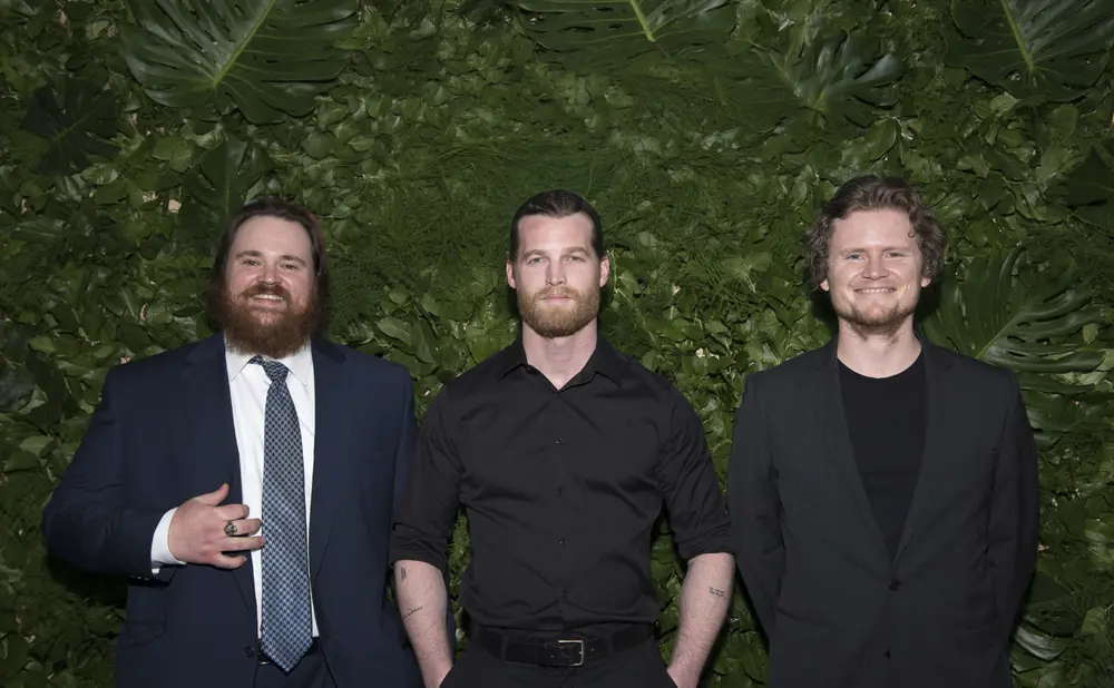 Three men in suits stand smiling in front of a green, leafy backdrop.