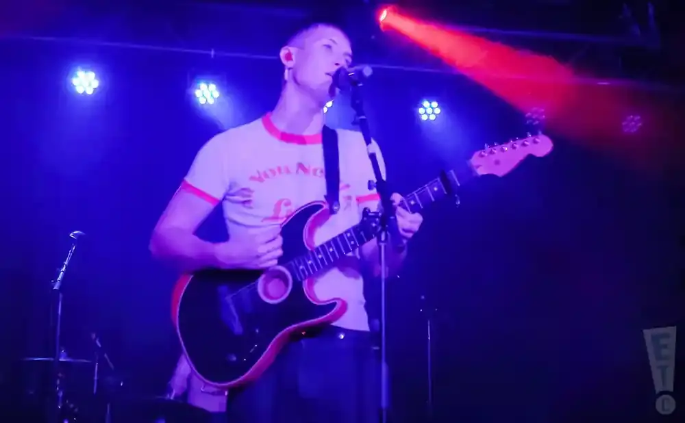 A male guitarist sings into a microphone while playing a black electric guitar under blue and red stage lights.