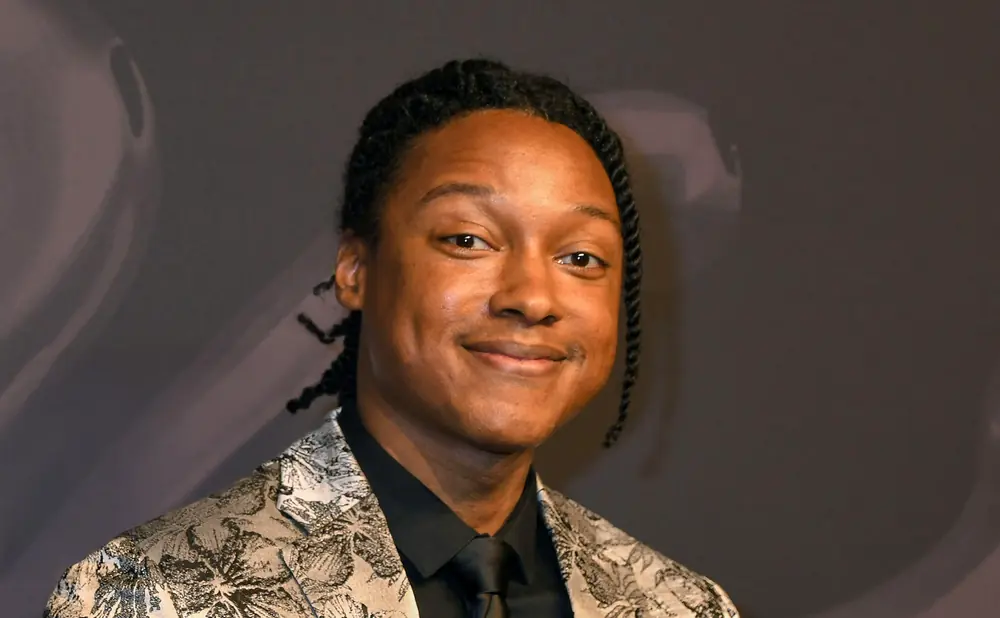 A Black man with braided hair wears a patterned suit and a black shirt and tie, smiling at the camera.