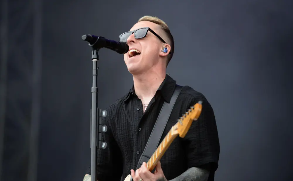 Musician with sunglasses and tattoos singing into a microphone while playing a guitar.