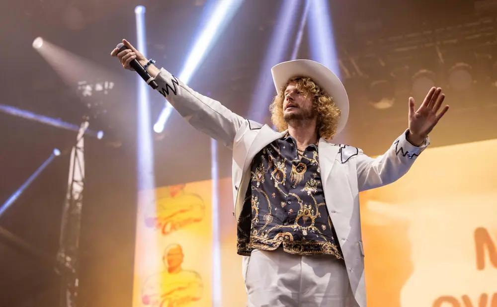 A male singer in a white cowboy hat and suit performs on stage with lights and a banner behind him.