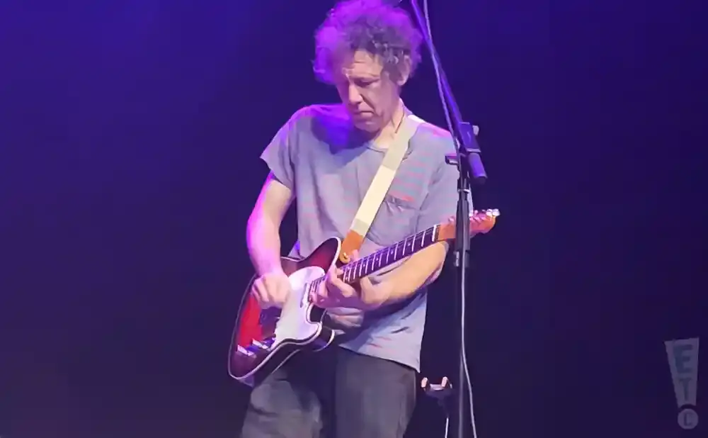 A guitarist with curly hair plays a red and white electric guitar on a dark stage with purple lighting.