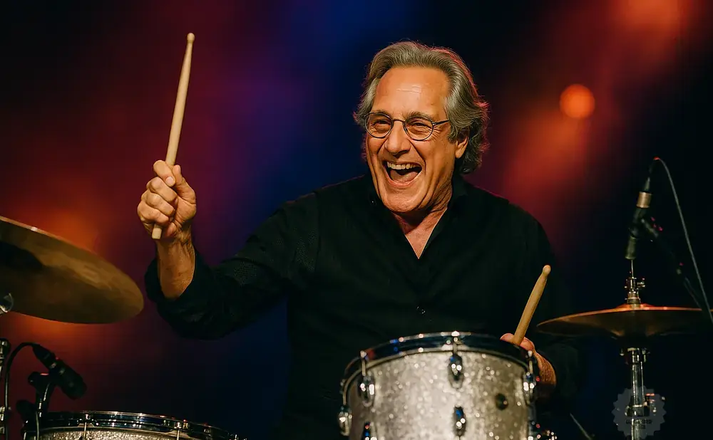 A laughing male drummer in a black shirt plays drums under colorful stage lights.