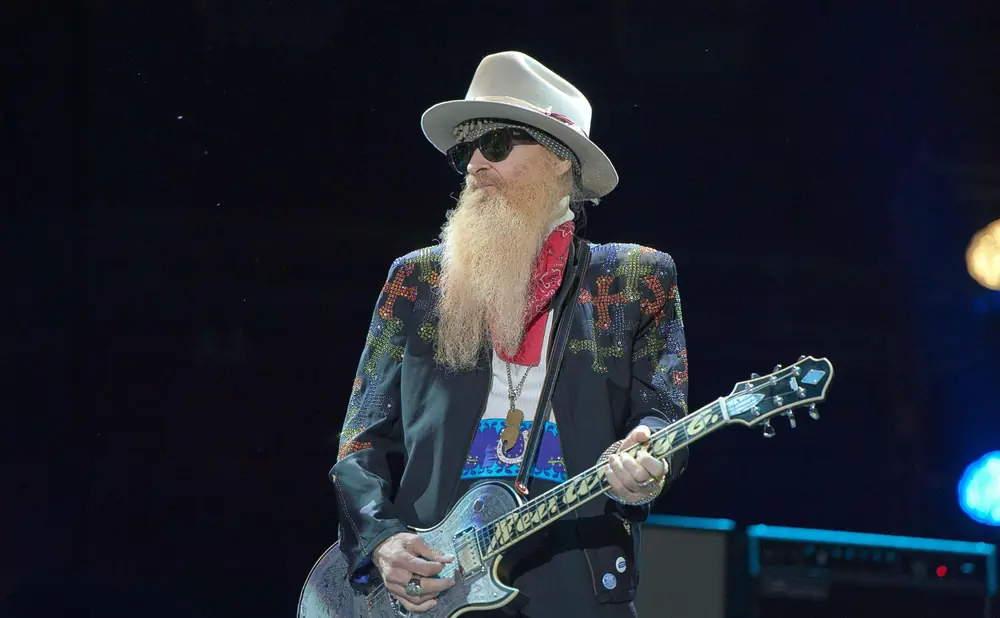 A male guitarist with a long blonde beard, wearing a hat and sunglasses, plays a decorated guitar on stage.