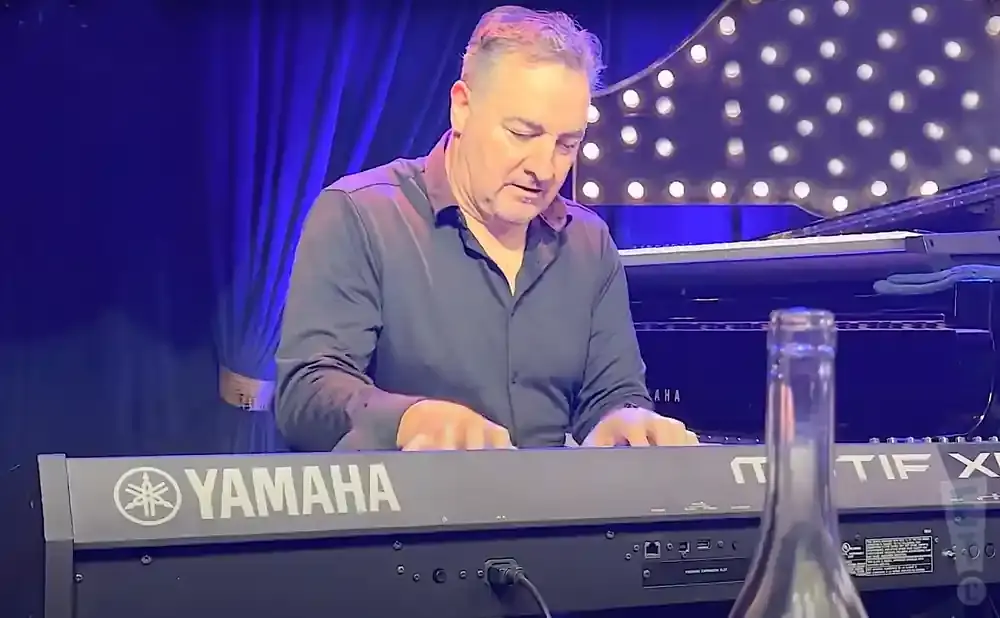 A man plays a Yamaha keyboard in front of a grand piano, with a blurred bottle in the foreground.