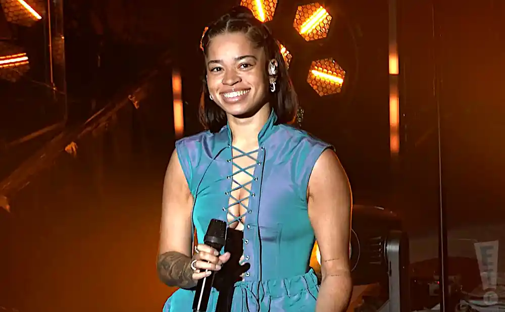 Singer Ella Mai smiles while performing on stage in a blue lace-up top and gold earrings.