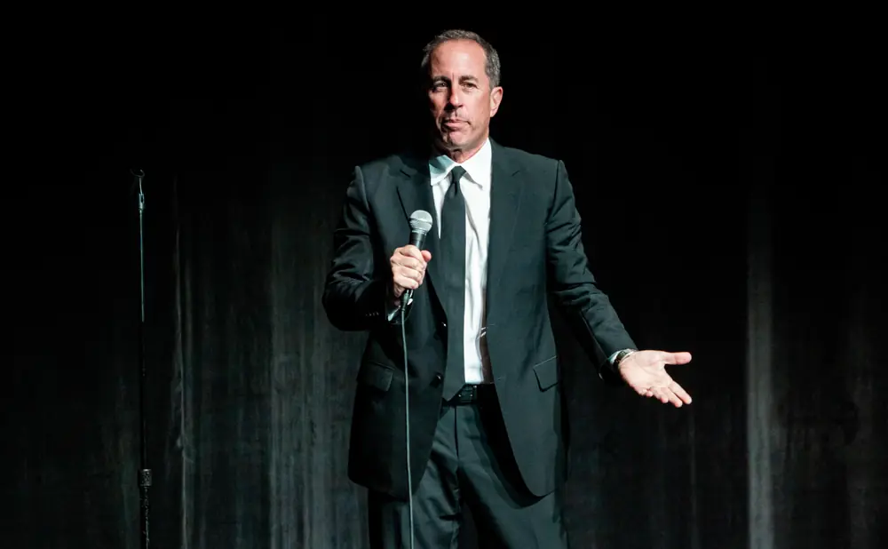 Jerry Seinfeld stands on stage in a black suit, holding a microphone and gesturing with his hand.