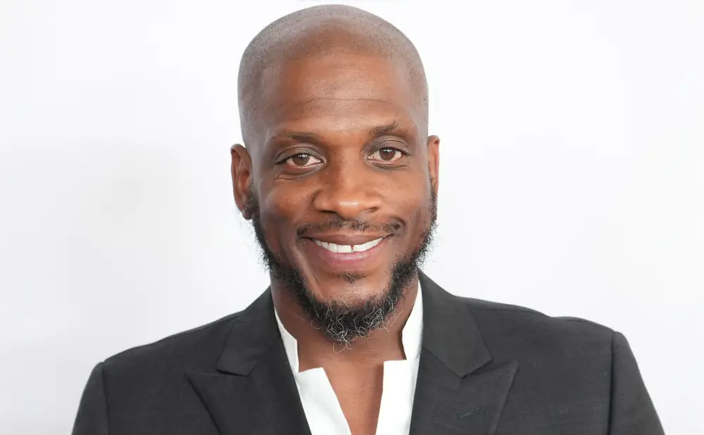 A bald Black man with a microphone in his hand, wearing a polo shirt and speaking with exaggerated expression.