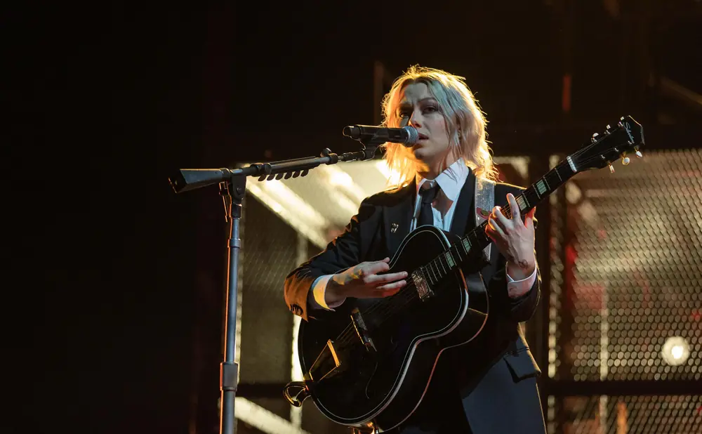 A woman with blonde hair plays a black guitar and sings into a microphone on stage.