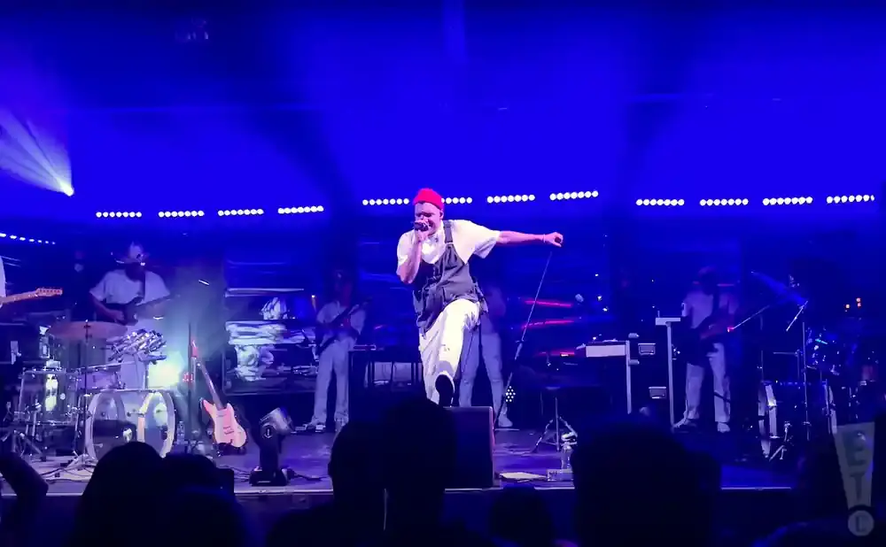 A male singer in a red hat and overalls performs on a brightly lit blue stage with a band.