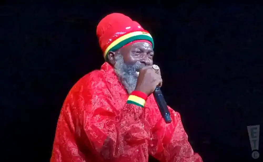 A man in a red outfit and hat with Jamaican colors sings into a microphone.