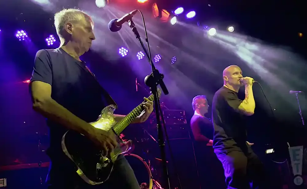 A bald man sings into a microphone on a stage, with a guitarist and another man behind him.