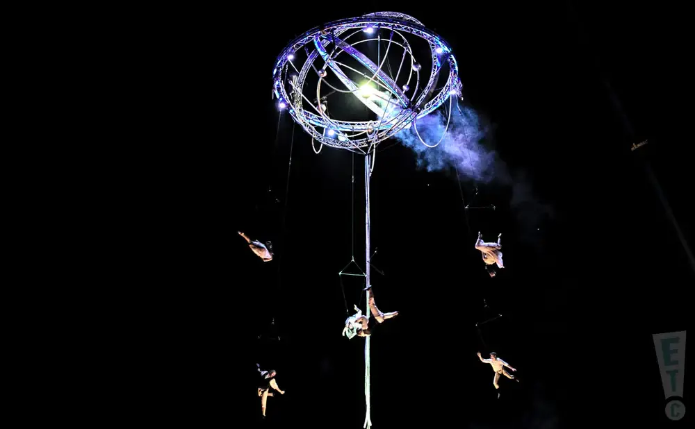 Aerial performers hang from a luminous, spherical apparatus against a black sky.