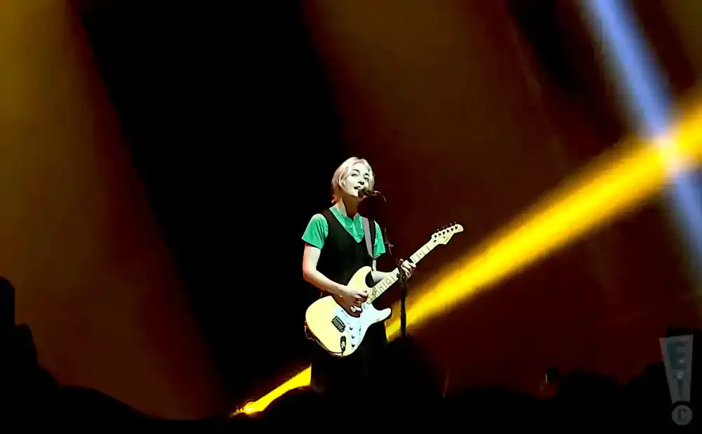 A person with short blonde hair plays a cream-colored electric guitar on a dark stage lit by yellow and blue spotlights.