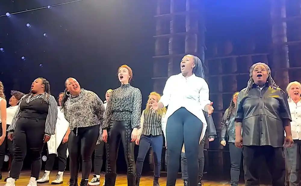 A choir of Black women and girls sing on a dimly lit stage.