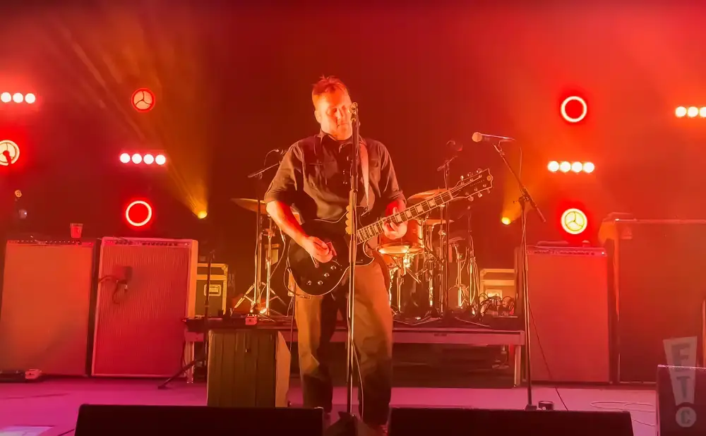 A man plays a black electric guitar on stage, bathed in red and orange stage lights.