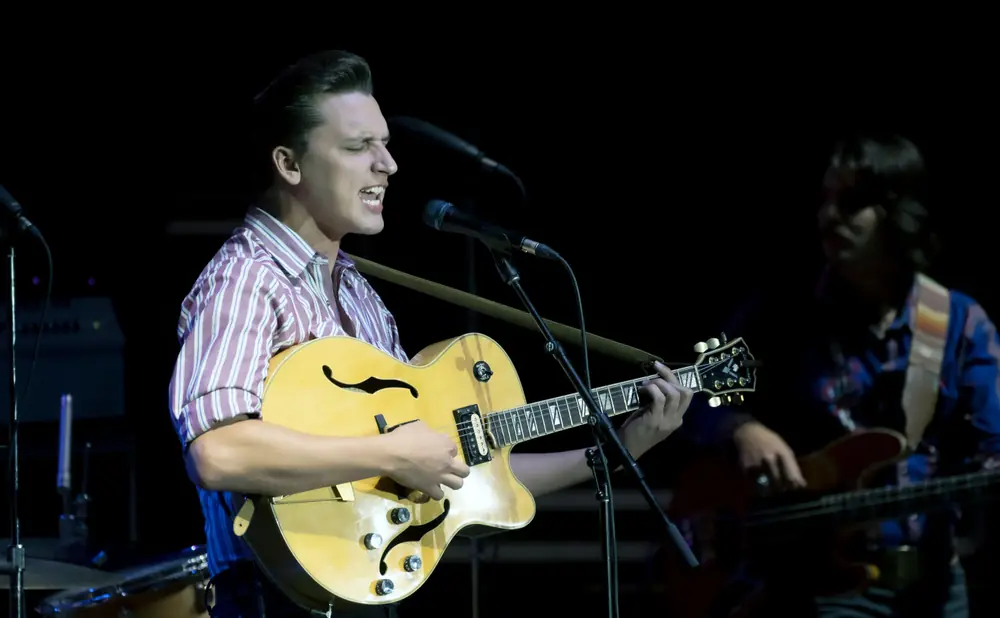 A man in a striped shirt sings and plays a hollow-body electric guitar on stage.