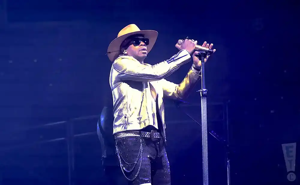 Man in a gold jacket and cowboy hat sings into a microphone on stage with blue lighting.