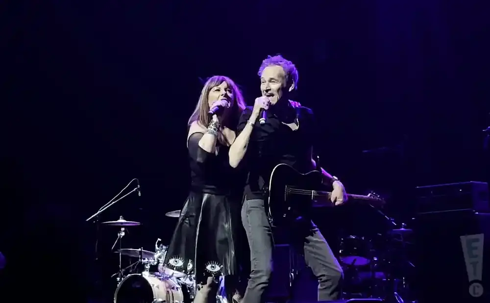 A man and woman sing into microphones on a dark stage with instruments.