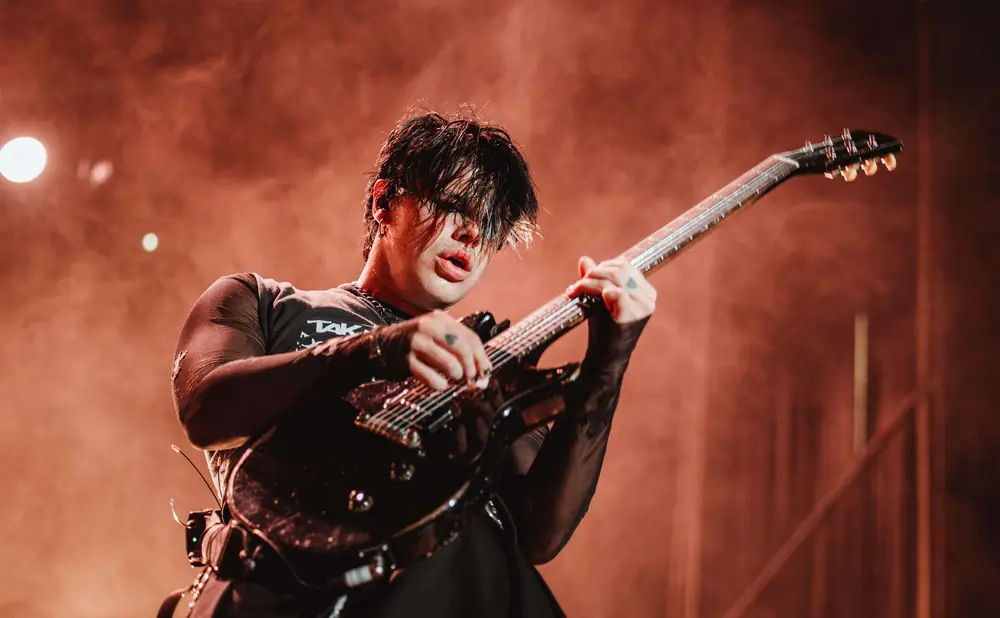 A musician with dark hair plays a black electric guitar on a stage, illuminated by warm, smoky lighting.