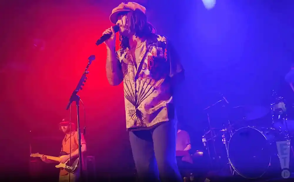 A man in a hat and patterned shirt sings into a microphone on a stage with red and blue lighting.