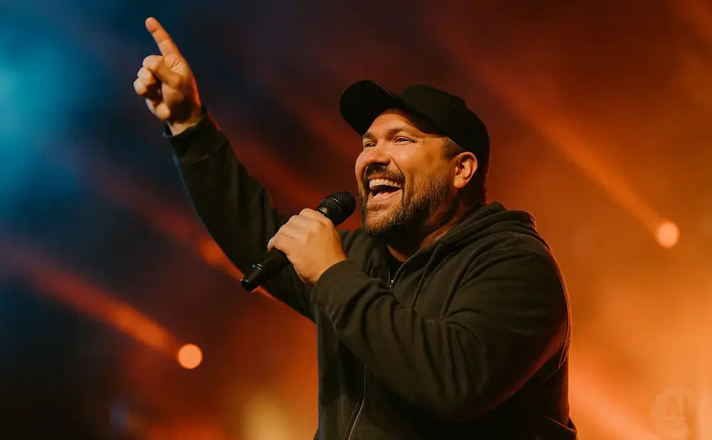A man in a baseball cap holds a microphone and points up, smiling and laughing on a stage with orange lighting.
