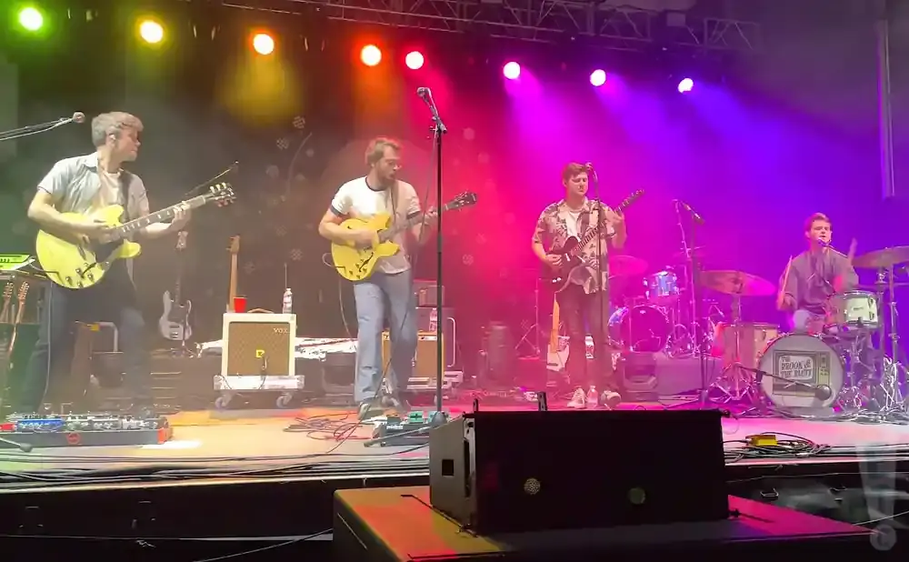 A band performs on a stage with purple and pink lighting. Two guitarists are on the left, and a drummer is on the right.