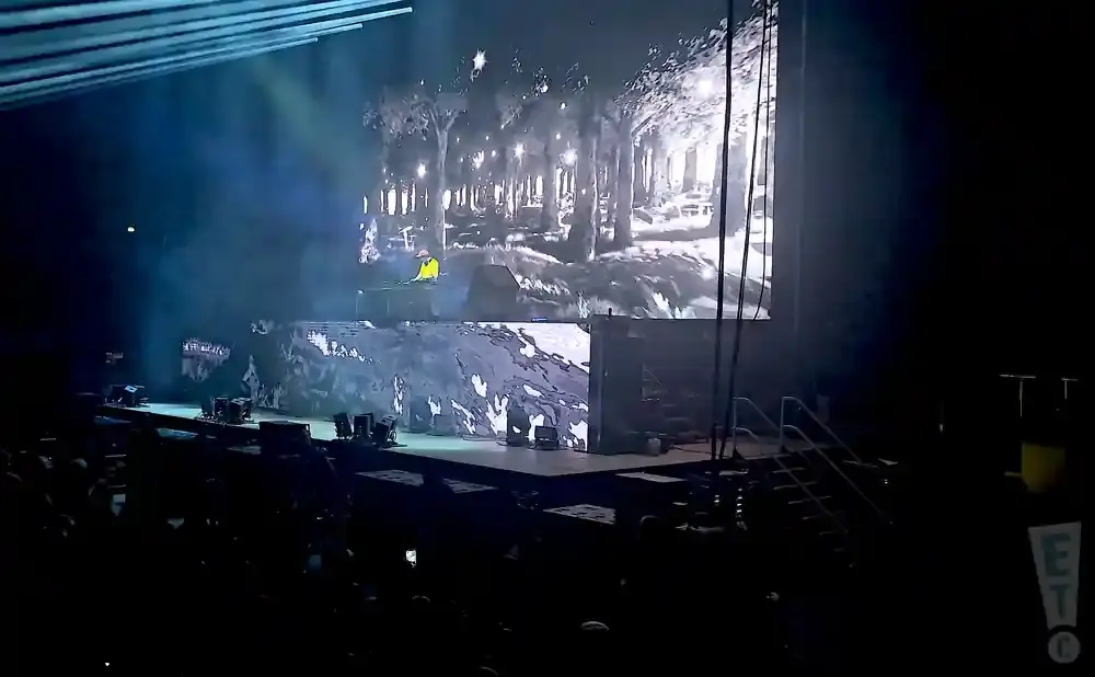 A performer in a yellow jacket stands at a DJ booth on a stage, with a large screen displaying a forest scene behind them.