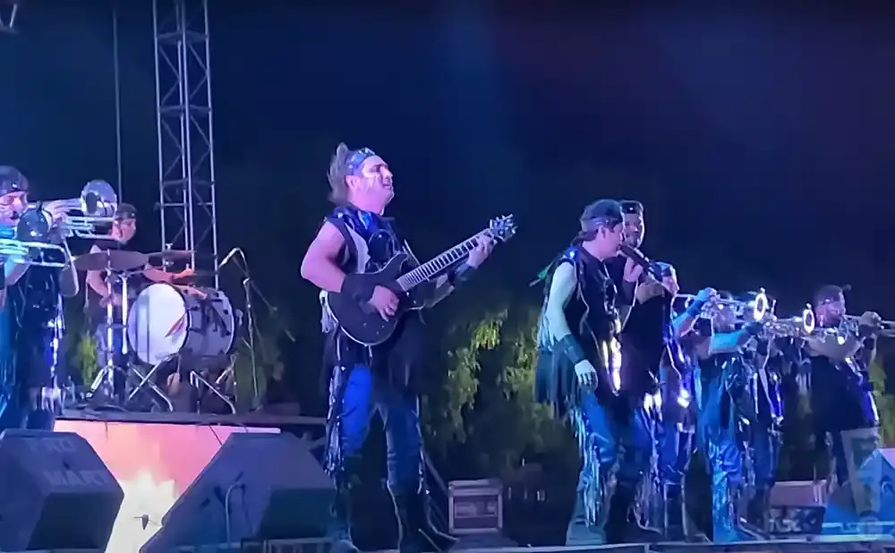A band performs on stage at night. A guitarist sings while others play brass instruments and drums.