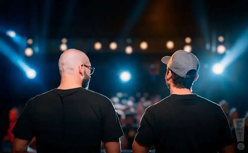 Two men, one bald with glasses and the other wearing a baseball cap, stand with their backs to the camera facing a stage with bright lights.