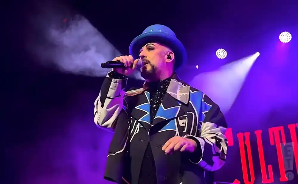 Boy George sings into a microphone on stage, wearing a blue hat and a colorful jacket under purple lights.
