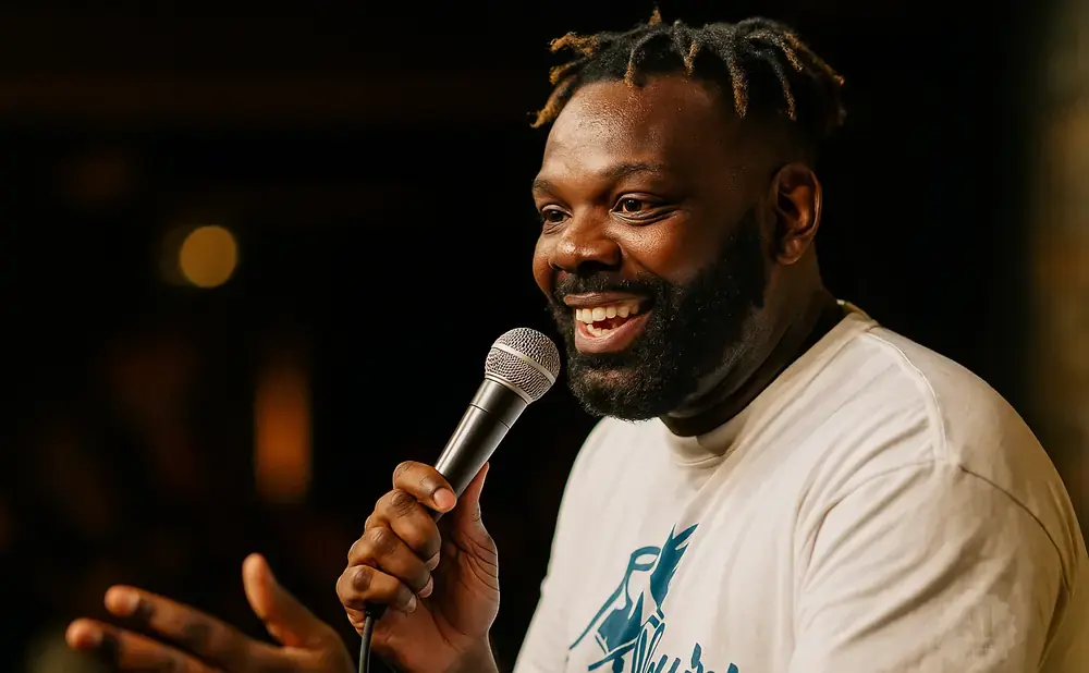 A Black man with dreadlocks and a beard laughs while holding a microphone and gesturing with his hand.