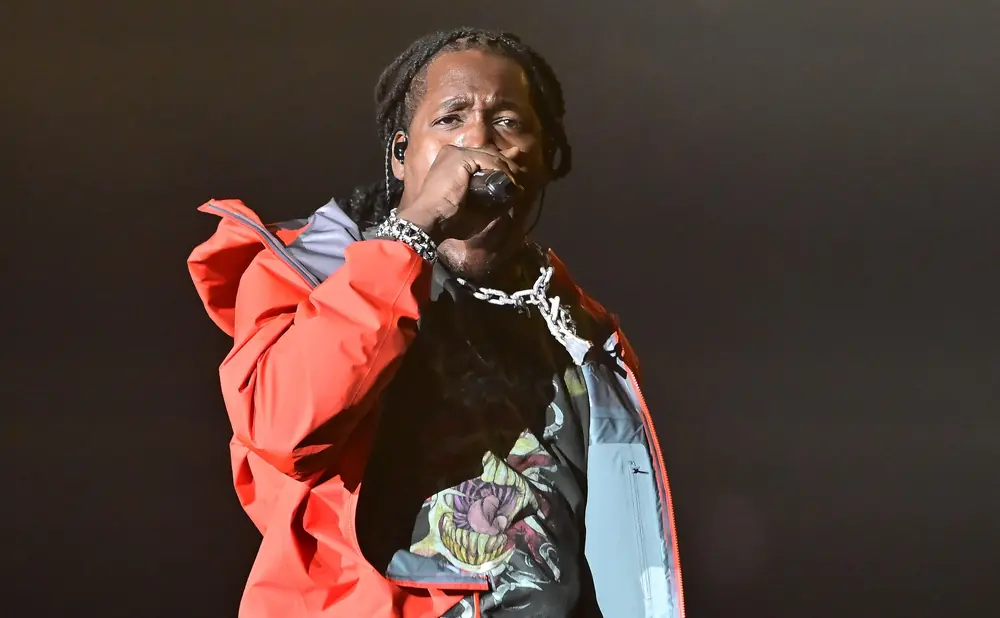 A rapper with dreadlocks, wearing a red jacket and a chain, performs into a microphone.