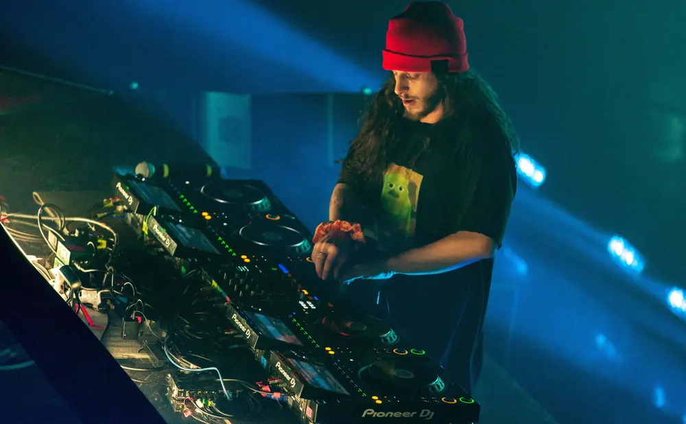 DJ with long dark hair and red beanie, performing on Pioneer DJ equipment under blue stage lights.