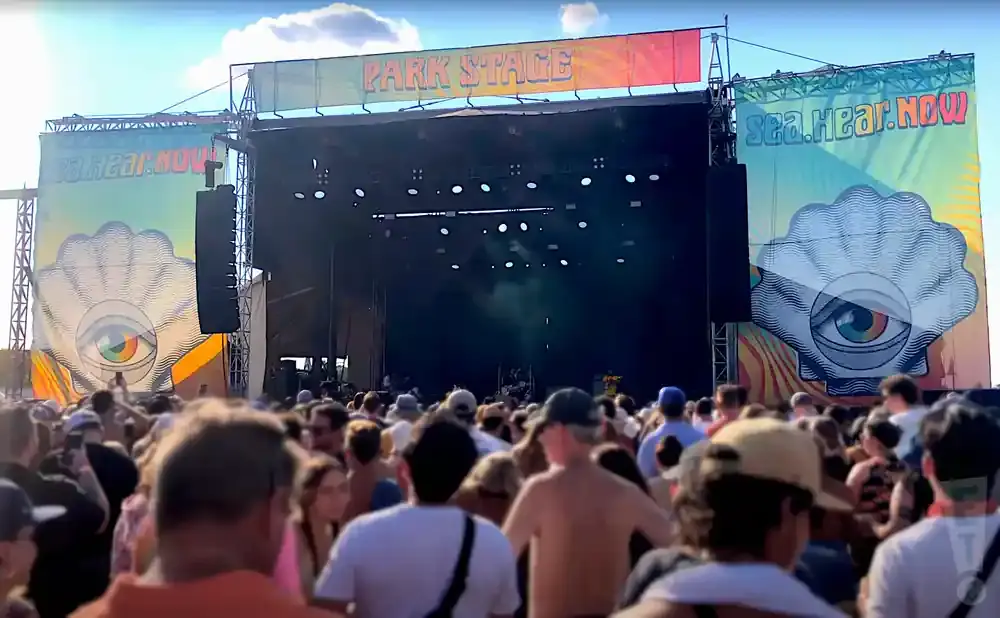 A crowded outdoor concert stage with banners, lights, and a large shell graphic with an eye.