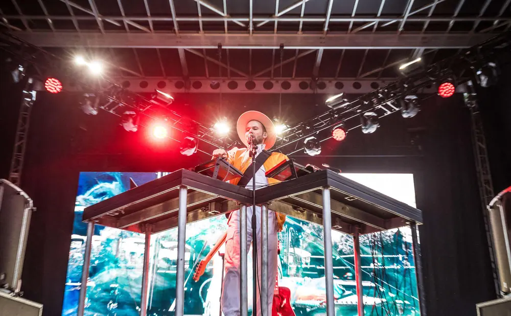 Musician in a hat and orange jacket plays a keyboard on stage with a large screen behind him.